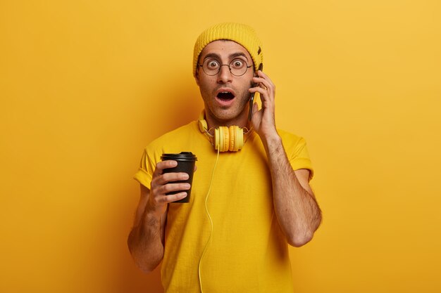 Un joven sorprendido tiene una conversación telefónica, jadea nerviosamente, mira con pánico, usa una camiseta informal, un sombrero y gafas