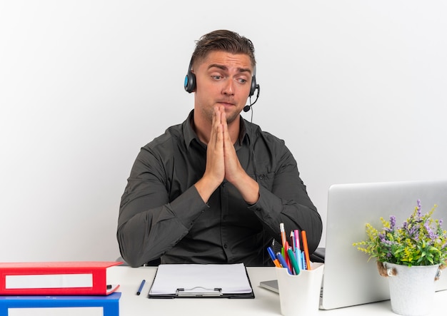 Joven sorprendido trabajador de oficina rubia en auriculares se sienta en el escritorio con herramientas de oficina mirando portátil sostiene las manos juntas aisladas sobre fondo blanco con espacio de copia