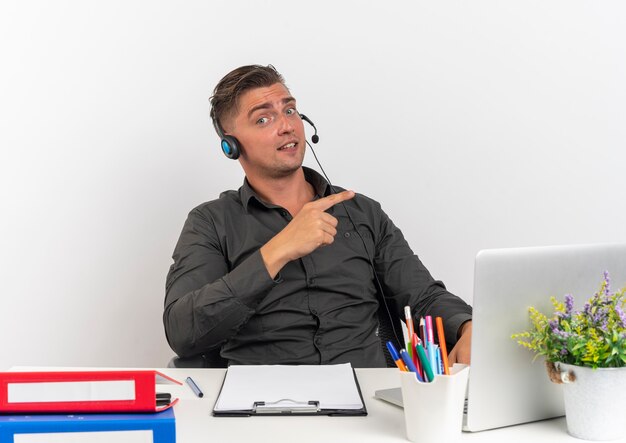 Joven sorprendido trabajador de oficina rubia en auriculares se sienta en el escritorio con herramientas de oficina usando puntos de portátil en el lado aislado sobre fondo blanco con espacio de copia