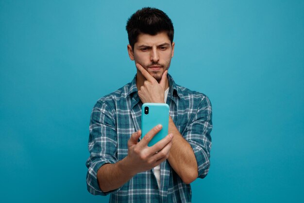 Joven sospechoso manteniendo la mano en la barbilla estirando el teléfono móvil hacia la cámara mirándolo aislado en el fondo azul