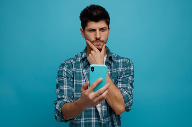 Joven sospechoso manteniendo la mano en la barbilla estirando el teléfono móvil hacia la cámara mirándolo aislado en el fondo azul