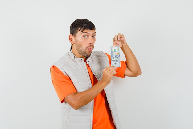 Joven sosteniendo billete de un dólar en camiseta, chaqueta y mirando asombrado, vista frontal.