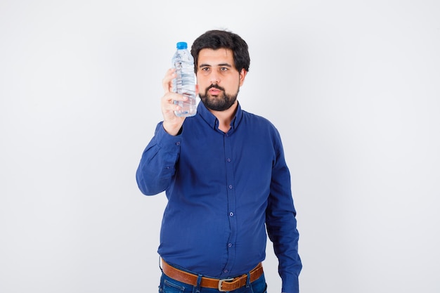 Joven sosteniendo una botella de agua y mirándolo con camisa azul y jeans y mirando serio. vista frontal.