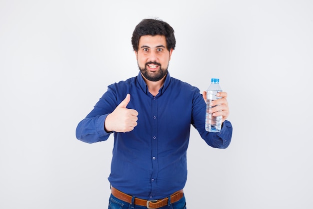 Joven sosteniendo una botella de agua y mostrando el pulgar hacia arriba en camisa azul y jeans y luciendo optimista. vista frontal.