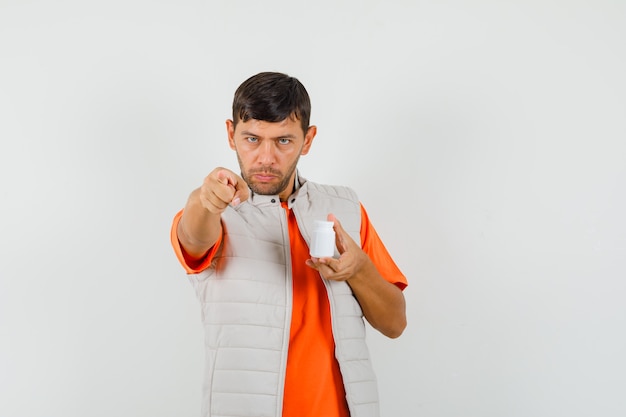 Joven sosteniendo una botella de píldoras, apuntando a la cámara en camiseta, chaqueta y mirando confiado. vista frontal.