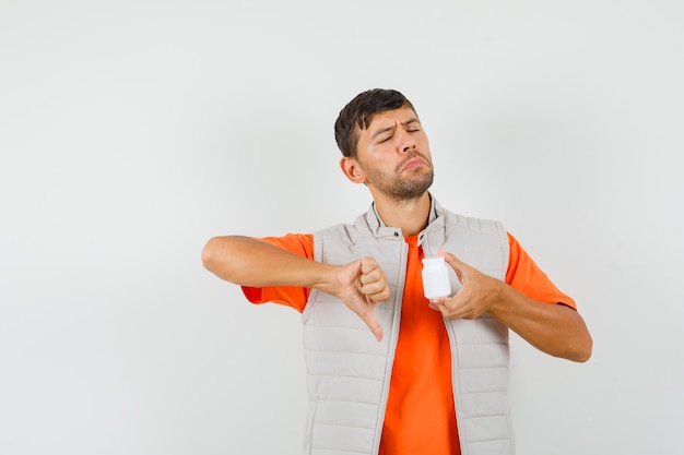 Joven sosteniendo una botella de píldoras, mostrando el pulgar hacia abajo en camiseta, chaqueta y mirando descontento. vista frontal.