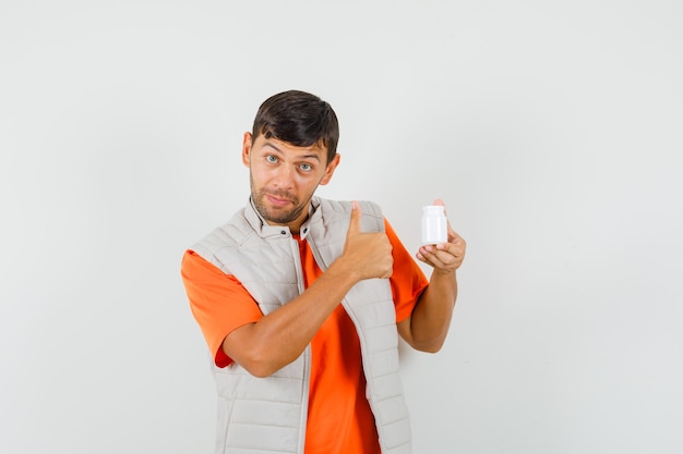 Foto gratuita joven sosteniendo una botella de píldoras, mostrando el pulgar hacia arriba en camiseta, chaqueta y mirando satisfecho. vista frontal.