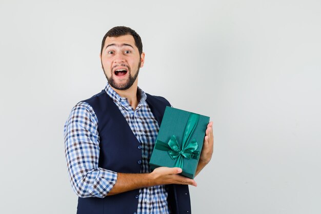 Joven sosteniendo la caja actual en camisa, chaleco y mirando contento.