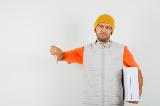 Foto gratuita joven sosteniendo una caja de cartón, mostrando el pulgar hacia abajo en camiseta, chaqueta, sombrero y mirando disgustado, vista frontal.