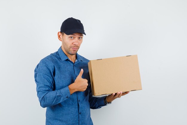 Joven sosteniendo una caja de cartón con el pulgar hacia arriba en camisa azul, gorra y mirando optimista, vista frontal.