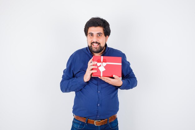 Joven sosteniendo la caja de regalo con ambas manos en camisa azul y jeans y mirando optimista, vista frontal.