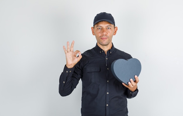 Joven sosteniendo caja de regalo y haciendo bien firmar en camisa negra con gorra