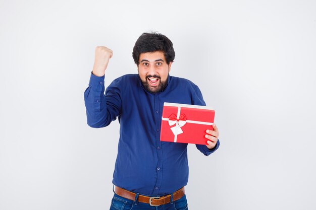 Joven sosteniendo una caja de regalo y mostrando la pose de ganador en camisa azul y jeans y luciendo optimista, vista frontal.