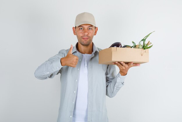 Joven sosteniendo la caja de verduras con el pulgar hacia arriba en camisa con gorra y mirando positivo