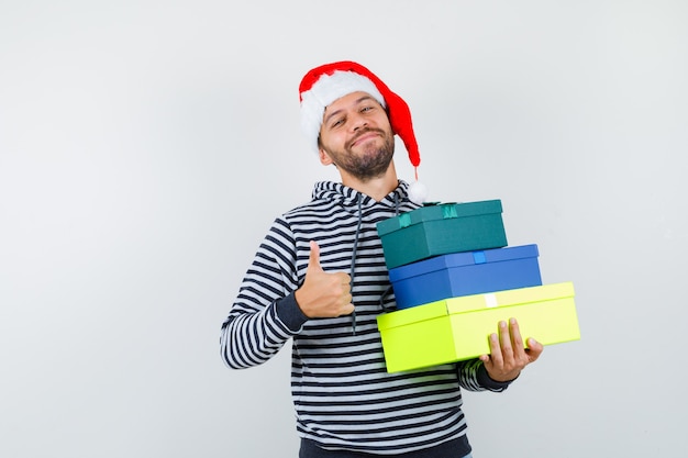 Joven sosteniendo cajas presentes, mostrando el pulgar hacia arriba en sudadera con capucha, gorro de Papá Noel y luciendo alegre.