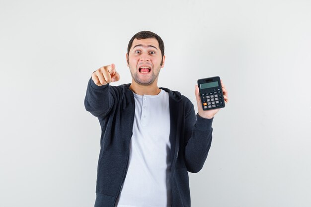 Joven sosteniendo la calculadora y apuntando con el dedo índice a la cámara en camiseta blanca y sudadera con capucha negra con cremallera frontal y mirando optimista, vista frontal.