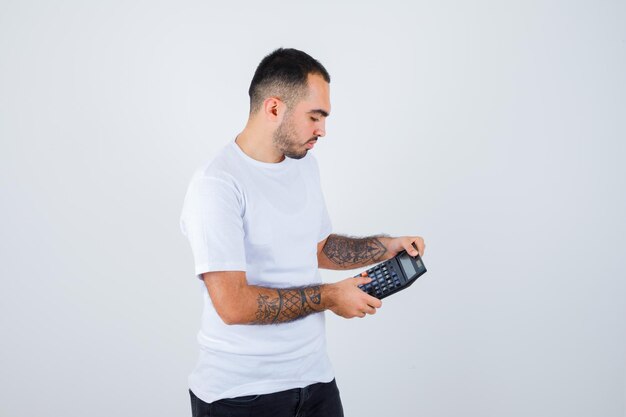 Joven sosteniendo la calculadora y haciendo operaciones en ella con camiseta blanca y pantalón negro y mirando serio