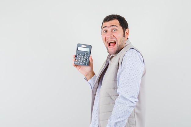 Joven sosteniendo la calculadora en una mano con chaqueta beige y gorra y mirando sorprendido, vista frontal.