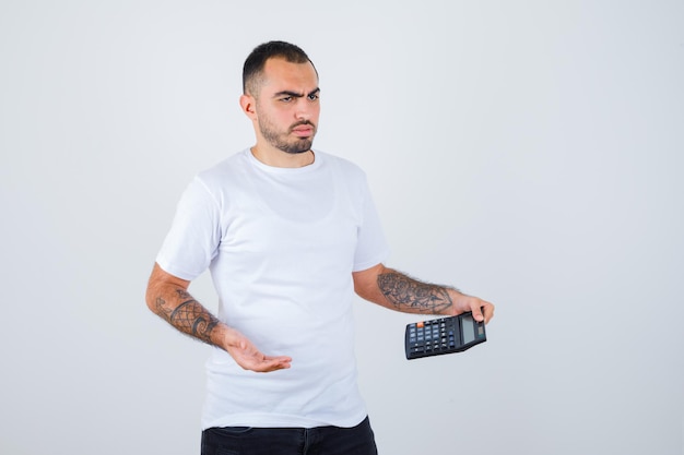 Joven sosteniendo calculadora y pensando en algo en camiseta blanca y pantalón negro y mirando pensativo