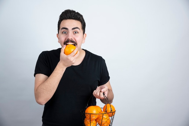 joven sosteniendo una canasta metálica llena de frutas y comiendo naranja.