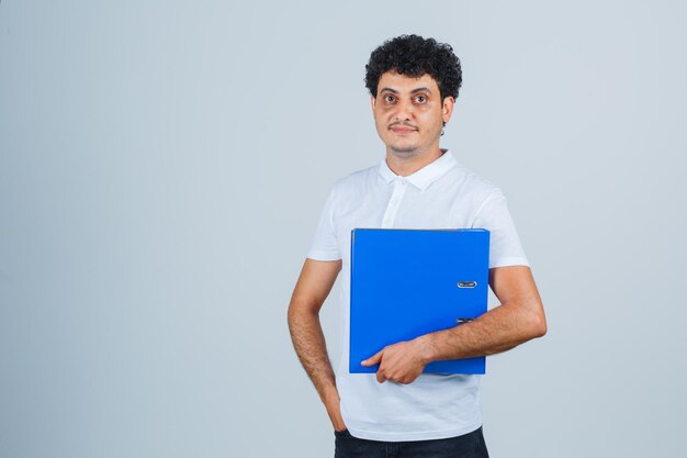 Joven sosteniendo la carpeta de archivos en camiseta blanca y jeans y mirando serio, vista frontal.