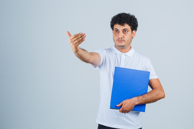 Joven sosteniendo la carpeta de archivos e invitando a venir con camiseta blanca y jeans y mirando serio. vista frontal.