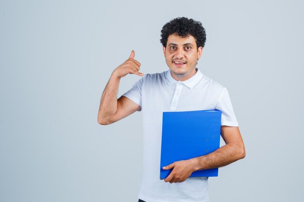 Joven sosteniendo la carpeta de archivos y mostrando el gesto de llamarme en camiseta blanca y jeans y mirando serio, vista frontal.