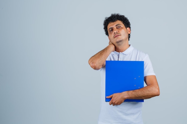 Joven sosteniendo la carpeta de archivos, sosteniendo la mano detrás del cuello en camiseta blanca y jeans y con aspecto cansado. vista frontal.