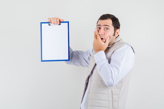 Joven sosteniendo el cuaderno y apuntando a él con chaqueta beige y mirando sorprendido, vista frontal.