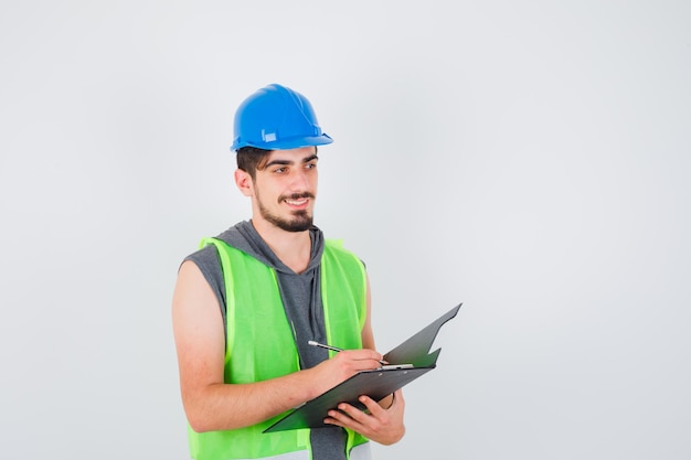 Joven sosteniendo el cuaderno y escribiendo algo en él con lápiz en uniforme de construcción y mirando feliz