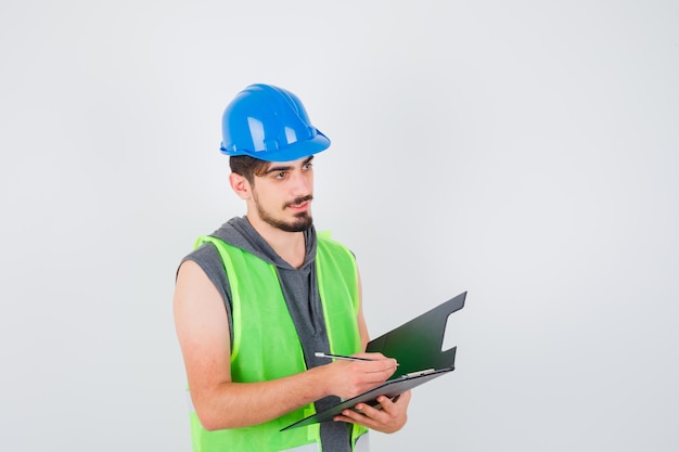 Joven sosteniendo el cuaderno y escribiendo algo en él con lápiz en uniforme de construcción y mirando pensativo