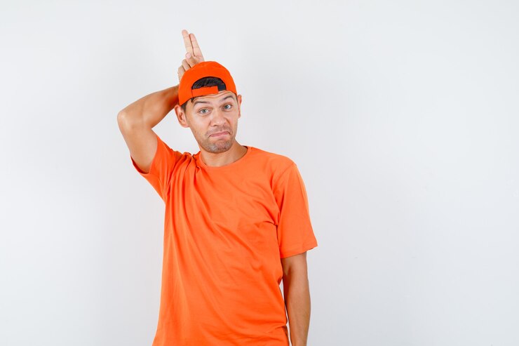 Foto gratuito joven sosteniendo dos dedos detrás de la cabeza como cuerno en camiseta naranja y gorra y mirando divertido