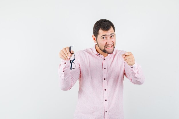 Joven sosteniendo gafas mientras apunta a la cámara con camisa rosa y mirando enojado