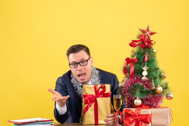 joven sosteniendo giftbox sentado en la mesa cerca del árbol de navidad y presenta en amarillo