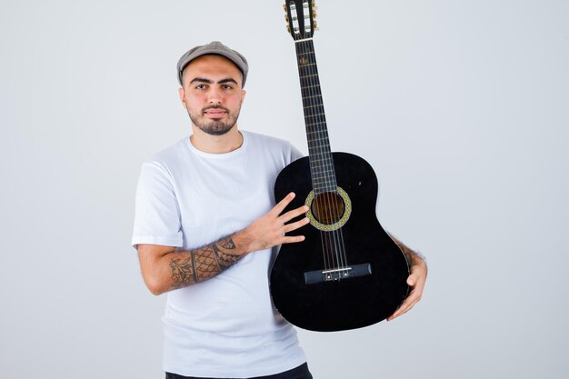 Joven sosteniendo la guitarra en camiseta blanca, pantalón negro, gorra gris y mirando serio