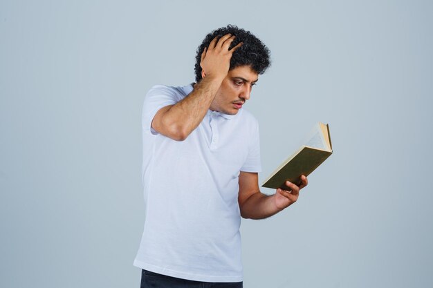 Joven sosteniendo el libro y rascándose la cabeza en camiseta blanca y jeans y mirando pensativo, vista frontal.