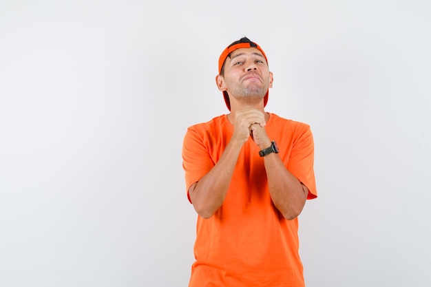 Joven sosteniendo las manos juntas en gesto de oración en camiseta naranja y gorra y con aspecto optimista