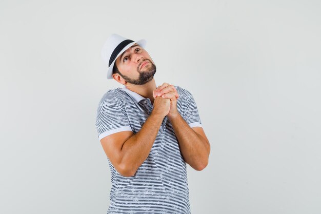 Joven sosteniendo las manos juntas en gesto de oración en camiseta, sombrero y mirando esperanzado. vista frontal.