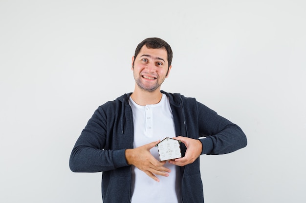 Joven sosteniendo el modelo de la casa con ambas manos y sonriendo en camiseta blanca y sudadera con capucha negra con cremallera frontal y con aspecto optimista. vista frontal.