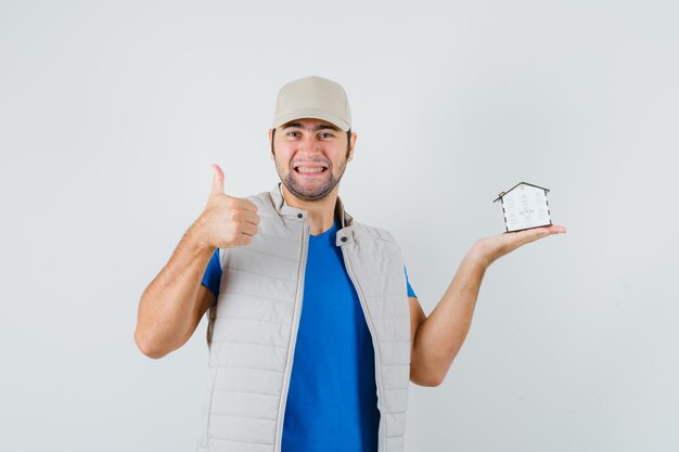 Joven sosteniendo el modelo de la casa, mostrando el pulgar hacia arriba en camiseta, chaqueta, gorra y mirando alegre. vista frontal.