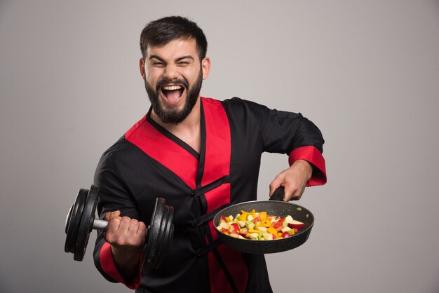 Joven sosteniendo una pesa y una sartén con verduras.