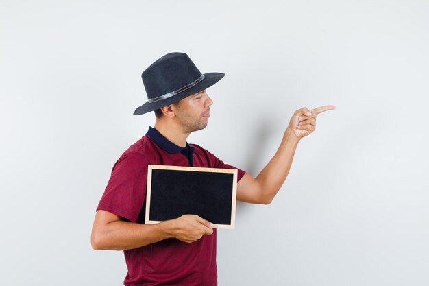 Joven sosteniendo la pizarra y apuntando al lado de la camiseta, vista frontal del sombrero.