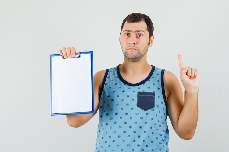 Foto gratuito joven sosteniendo el portapapeles, apuntando hacia arriba en camiseta azul y mirando desconcertado.