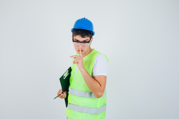 Joven sosteniendo portapapeles y bolígrafo, mostrando gesto de silencio en uniforme de construcción y mirando serio