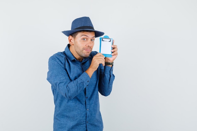 Joven sosteniendo el portapapeles con camisa azul, sombrero y mirando alegre, vista frontal.