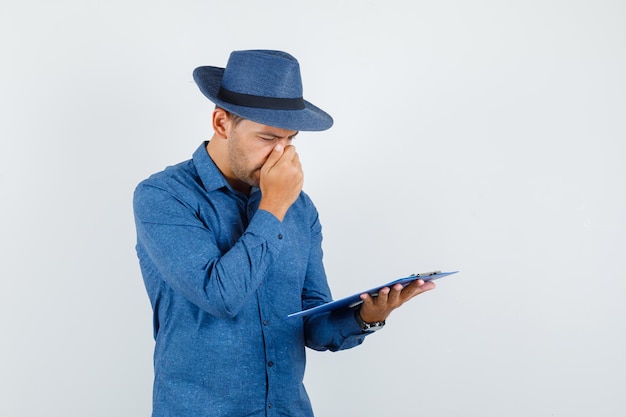 Foto gratuita joven sosteniendo el portapapeles con camisa azul, sombrero y mirando pensativo, vista frontal.