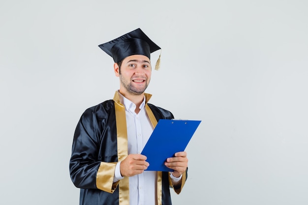 Joven sosteniendo el portapapeles en uniforme de posgrado y mirando alegre, vista frontal.