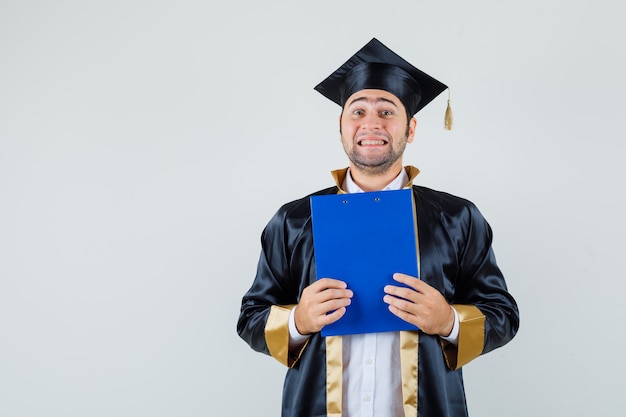 Foto gratuita joven sosteniendo el portapapeles en uniforme de posgrado y mirando alegre. vista frontal.