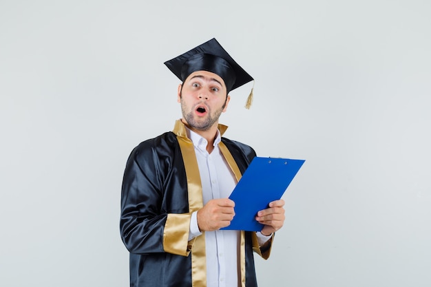 Foto gratuita joven sosteniendo el portapapeles en uniforme de posgrado y mirando sorprendido. vista frontal.