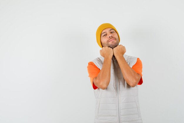 Joven sosteniendo los puños debajo de la barbilla en camiseta, chaqueta, sombrero y mirando alegre. vista frontal.